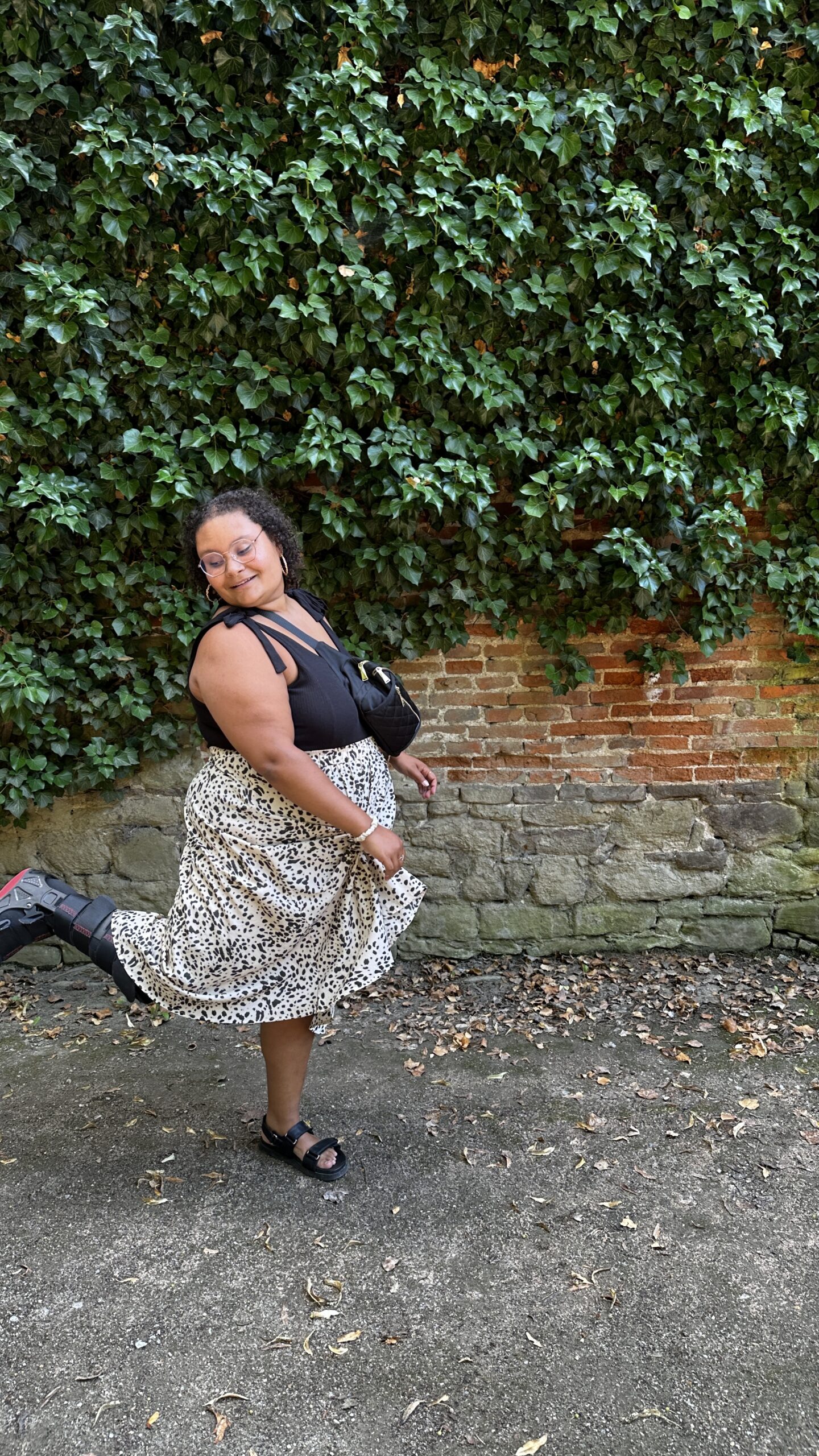 Ashley, a black woman with dark curly hair wearing gold framed glasses is twirling in her skirt and kicking up her walking boot foot. 