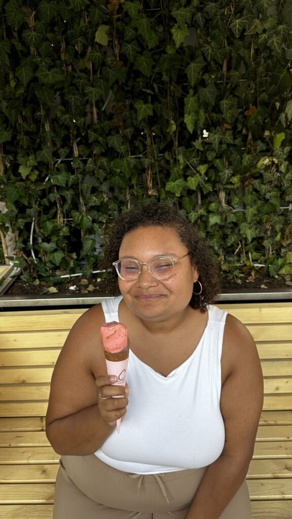 Ashley, a black woman with dark curly hair, wearing glasses and a white tank top holding gealto up and smiling.